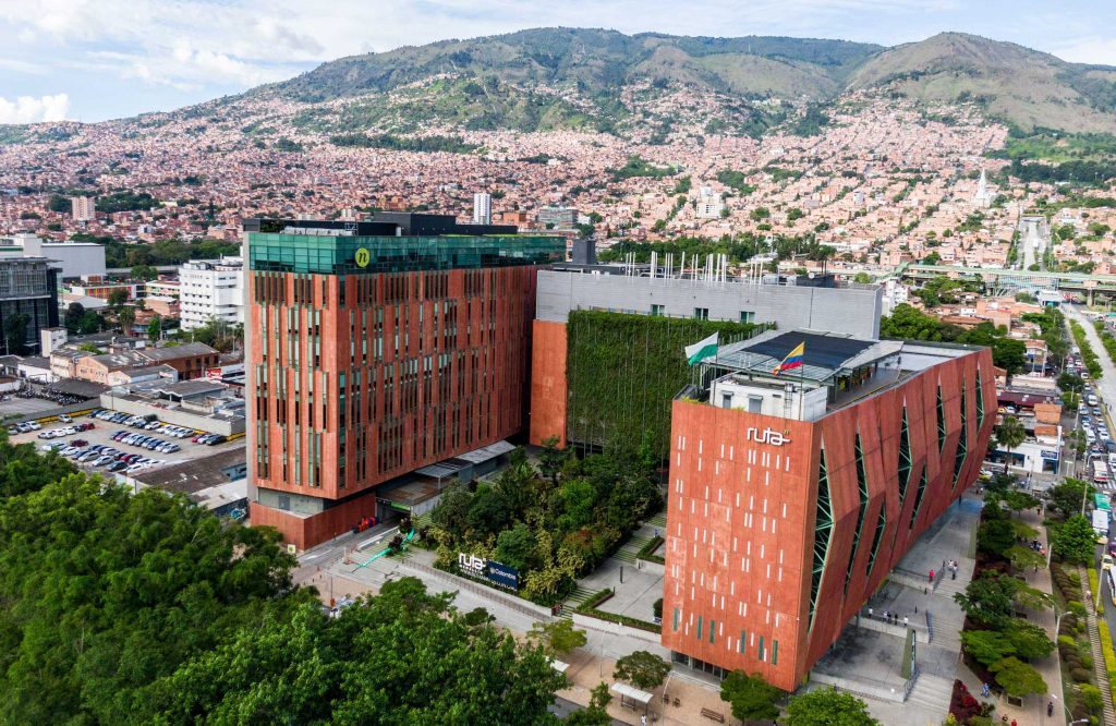 Aerial view of the Ruta N innovation district in Medellín — two modern rust-coloured buildings with green façades, set against the Aburrá Valley hillsides